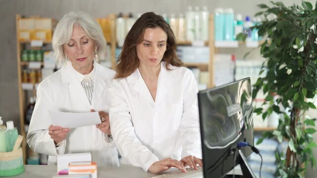 Senior female pharmacist helps a female assistant work with a computer in a pharmacy. They enter new arrivals of pills for head, migraine and allergy. Pharmacists work in a pharmacy . High quality 4k