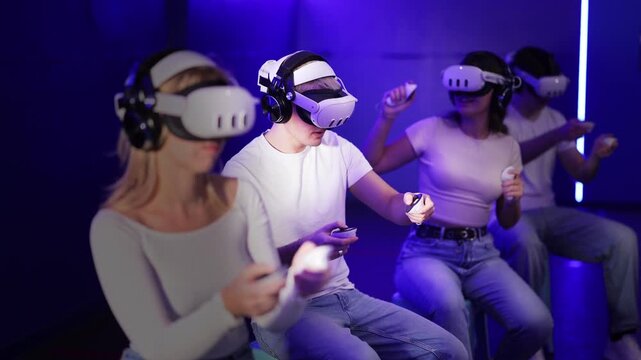 In darkened hall of gaming club, guy in VR helmet takes part in group game. Player overcomes obstacles and reaches goal in parallel reality. Imaginary fantastic world 