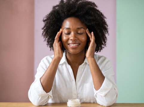 Relaxed African American woman with eyes closed massaging her temples near skincare product
