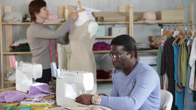 Experienced black middle-aged tailor stitching linen garment on sewing machine in workshop, while female colleague adjusting sample dress on mannequin 