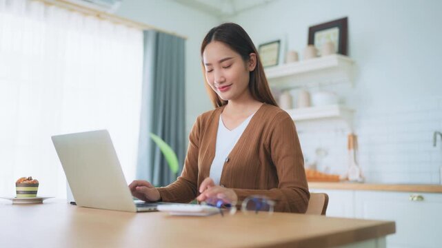 Beautiful young Asian woman working with laptop computer in home kitchen. Work at home