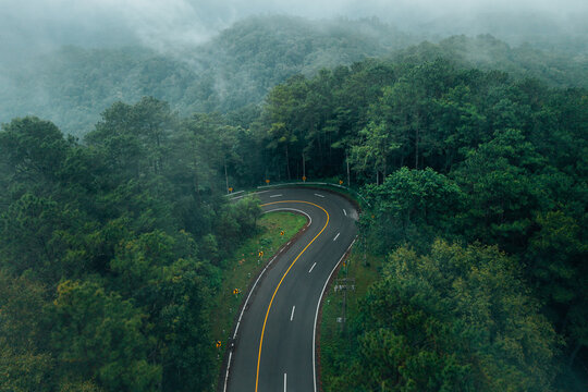 Atmospheric view of a winding road with yellow warning signs cutting through a dense misty pine forest on the scenic Chiang Mai to Pai route in Northern Thailand.
