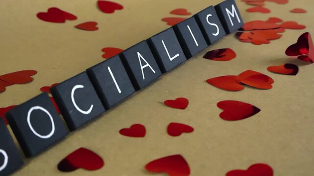 Camera slowly gliding over black tiles spelling the word socialism, surrounded by small, shiny red heart confetti on a light brown background, illustrating a love for the political ideology