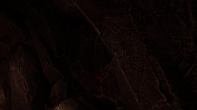 The scene shows a rocky surface inside a cave. The walls are dark and uneven with various textures. Shadows are visible due to the limited light. The environment feels enclosed and rugged.