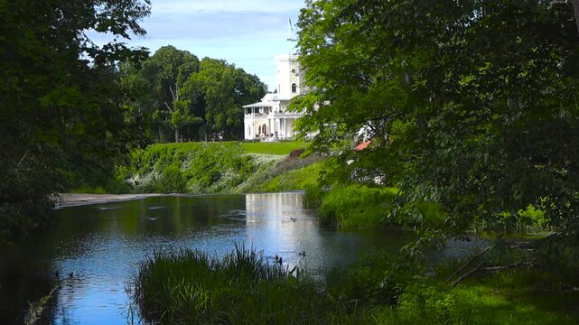Beautiful summer scenery with the Keila Joa manor reflected in the calm waters of the river, framed by lush green trees and reeds under a bright blue sky with some passing clouds