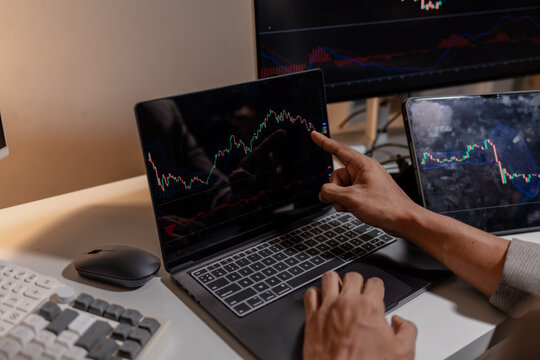 Professional trader analyzing stock market charts across multiple screens using laptop and tablet to monitor price movements, technical indicators and investment strategies in modern trading workspace
