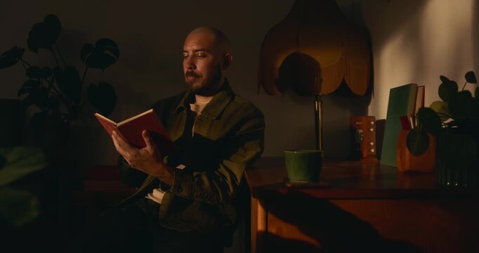 A relaxed bearded man with a shaved head reads a small red book at a wooden desk at night. The cozy room is dimly lit with warm light, plants, a vintage lamp, and a coffee mug in the background.