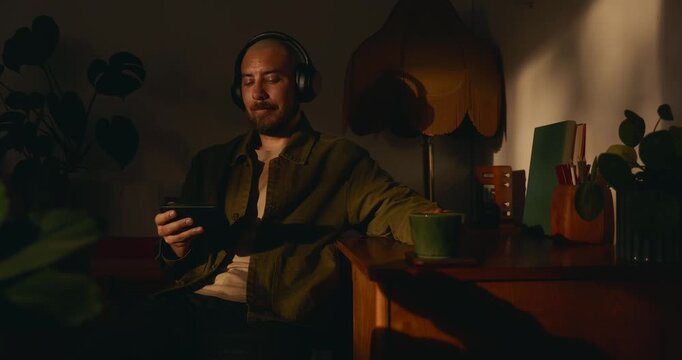 A relaxed adult man with headphones listens to music or a podcast while using his smartphone at a dimly lit cozy desk at home, enjoying a hot cup of coffee in warm evening lighting.