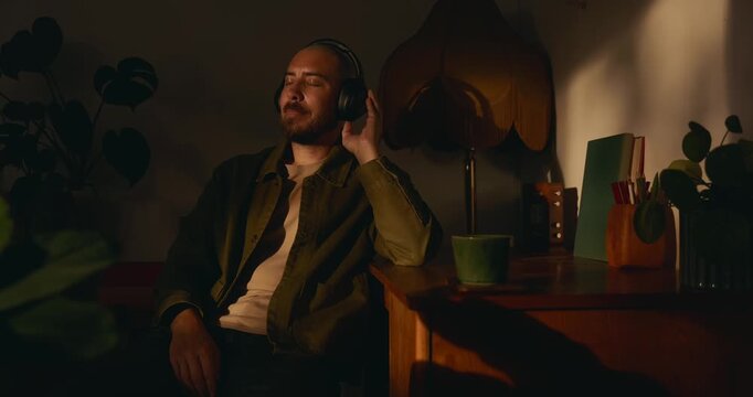 A peaceful bearded man sits at a desk with eyes closed, adjusting his wireless headphones to enjoy music or a podcast in a cozy, dimly warm-lit room with plants and a hot cup of coffee by his side.