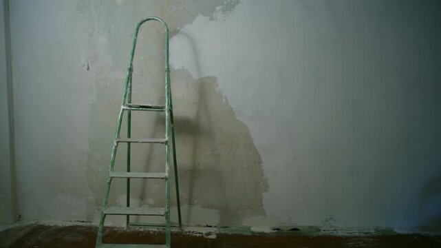 Green stepladder resting against a partially plastered wall in a room undergoing home improvement. The interior shows signs of recent repair work with drywall putty applied to the surface