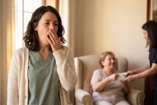 Caregiver yawning and covering mouth while an elder woman receives a cup of tea