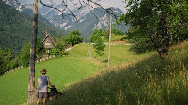 A farmer is mowing grass on a small alpine farm. The farm is located in a beautiful area with trees and mountains around. The sun shines bright as the farmer works hard.