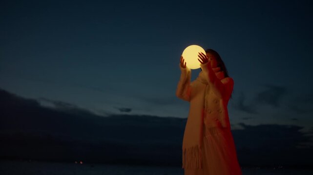 Young woman dressed warmly in scaf and formal clothes raises a glowing orb toward the dark sky bringing it close to cheeks by the sea, conveying wonder and longing as she holds it with care