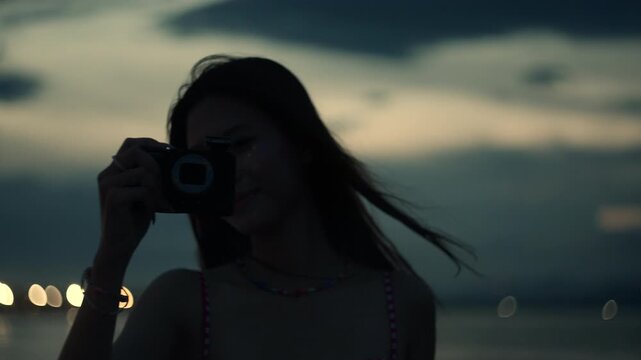 Woman taking a picture with a compact camera on the beach during blue hour, flash triggers illuminating the scene as she views the image smiling