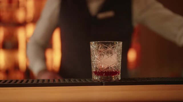 Bartender pouring red bitter cocktail into crystal glass with ice cube.