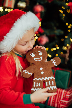 Girl wearing Santa hat holding gingerbread cookie sitting near decorated Christmas tree with gifts. Christmas and New Year holiday celebration concept