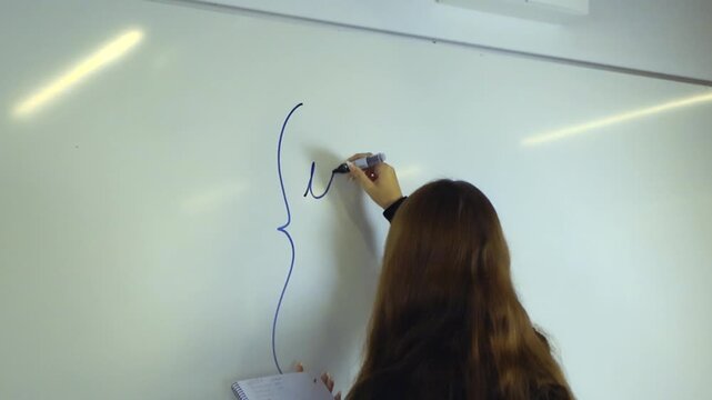 Female college student with long brown hair writing a complex mathematical formula on a classroom whiteboard with a blue marker, referencing notes from her spiral notebook during a lesson