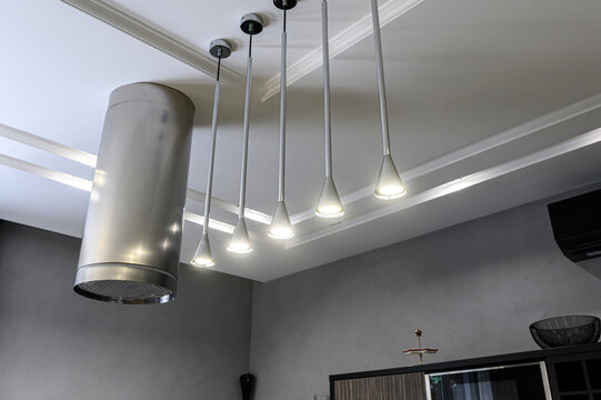 stainless‑steel cylindrical range hood sits beside sleek pendant lights with tapered shades. coffered ceiling, recessed lighting, muted gray walls define modern kitchen corner