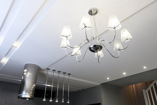 modern chandelier with curved chrome arms and frosted glass shades hangs from coffered ceiling. nearby, stainless‑steel range hood linear pendant lights accent sleek kitchen space