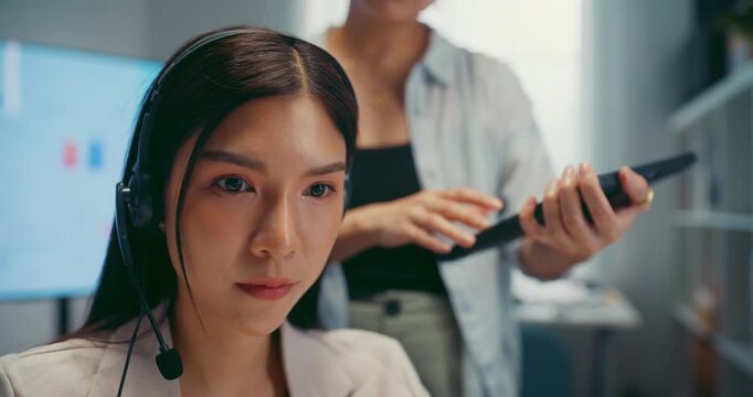 Young Asian businesswoman hold tablet to mentor trainee in call center workspace office. Corporate customer service training, employee support coach, telemarketing agent, teamwork help desk concept.