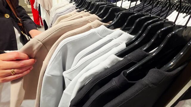 Customer hand selecting long-sleeved garments from clothing rack in department store, close-up view of merchandise in various neutral colors for fashion and retail concepts
