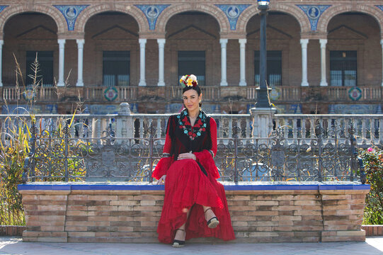 A beautiful flamenco dancer sitting on a tiled bench in a square in Seville, Spain. She is wearing a red ruffled dress and has a flower in her hair. Flamenco, a UNESCO Intangible Cultural Heritage.