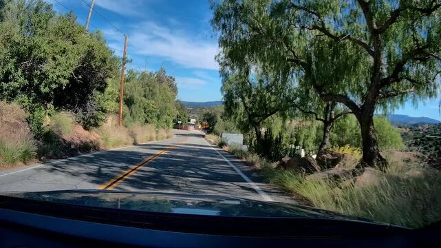 Dashcam view winding road drive through Santa Susanna Pass Road area towards Box Canyon Road, showing curve signs, trees, and houses under a bright blue sky with wispy clouds during daytime