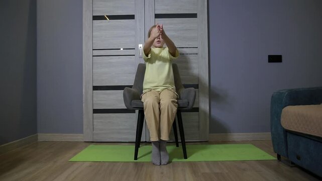 Young caucasian girl sits on chair performing arm exercises indoors, fitness activity for child, physical workout routine at home, promoting health and wellness for kids