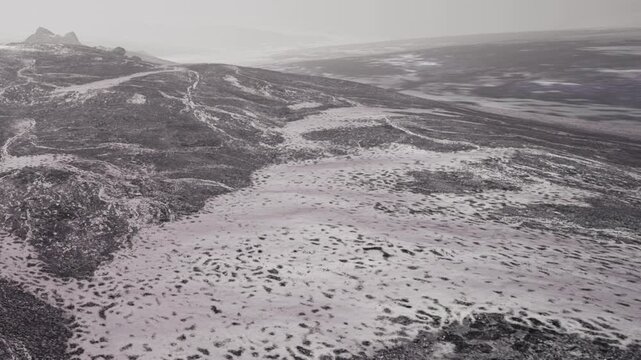 A wide view shows a snowy landscape with patches of dark rocks and distant hills. The scene captures the starkness of nature during winter, with no signs of life visible.