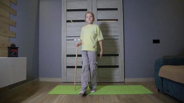Little girl exercising at home using stick for balance on green mat, performing light physical activity indoors, focusing on coordination and movement.
