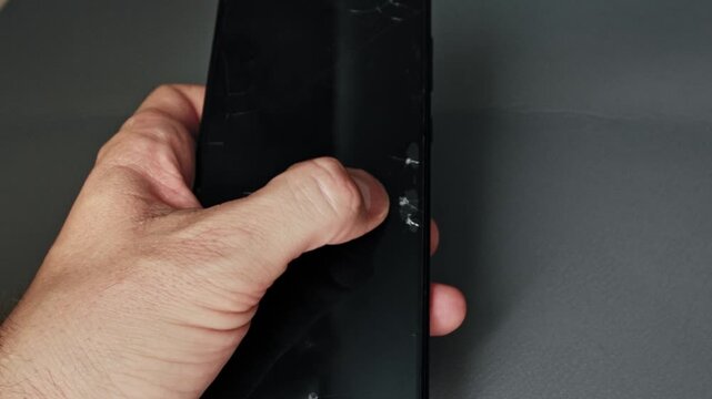 Male hand grips modern black smartphone with severely damaged cracked screen protector indicating accidental impact substantial wear over time with visible signs damage