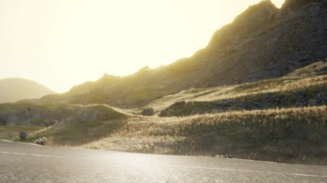 The sun rises over mountains in the background. A road runs through the landscape. Grass covers the ground near the road. The area looks quiet and natural.
