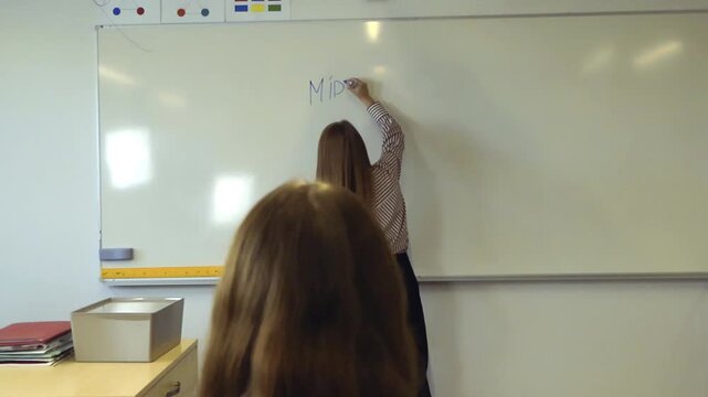 Female teacher writing the words Middle Eastern Conflict on a whiteboard during a lesson as a student watches from her desk, focusing on contemporary geopolitical issues and education