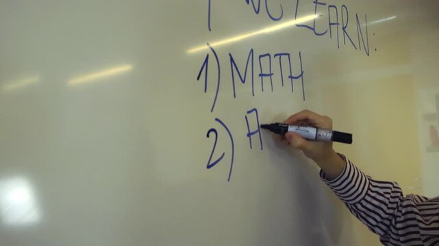 Professional educator's hand writing a lesson plan with a blue marker on a whiteboard during a lecture, listing subjects like math and algebra for students in a school or university class