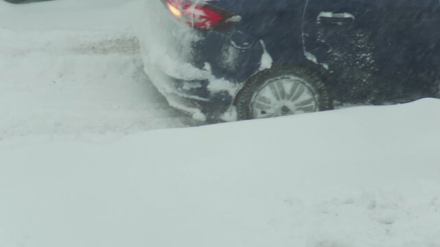 Dark blue car stuck in deep snow during winter blizzard, back wheel spinning on icy road conditions, emergency situation of automobile on snowy street, slow motion
