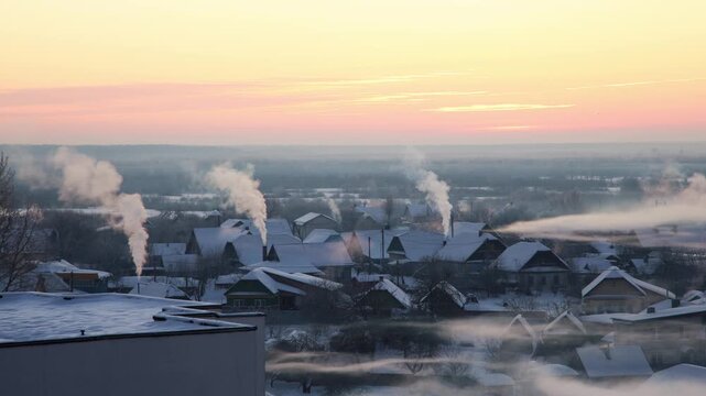 Winter village landscape at frosty morning sunrise with smoke rising from chimneys of snow-covered houses creating atmospheric scene