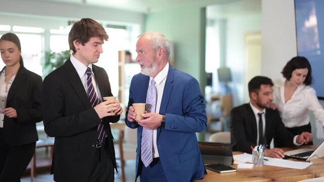 Elderly and adult male colleagues drinking coffee in office during break from work 