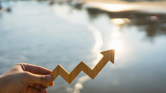 Economic growth and financial success concept with hand holding wooden upward trend arrow against glistening sea water and sunlight bokeh for stock market recovery and business profit.