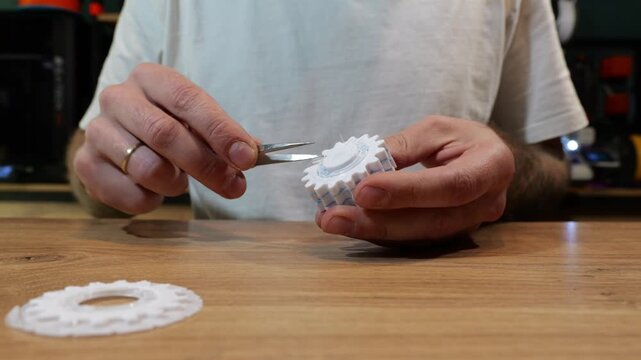 A man uses precision tweezers to remove blue PET-G support material from a white PLA 3D printed gear cog at a workshop table.