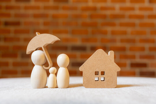 Family and home insurance concept with wooden peg doll figures under umbrella next to house model on table with brick background for property mortgage protection and financial security.