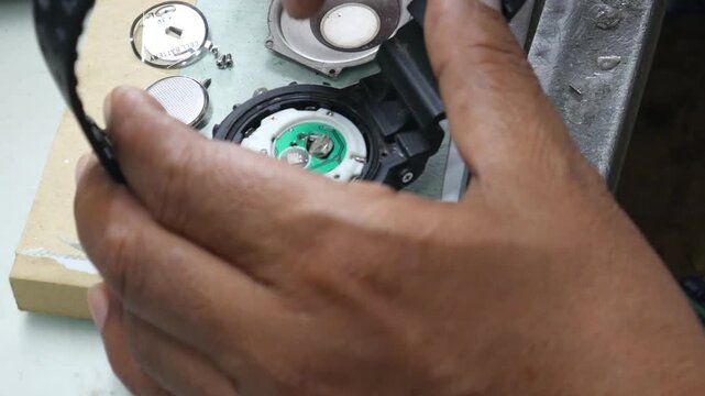 Watchmaker hands using tweezers and tools to repair digital watch movement on workbench in professional workshop.