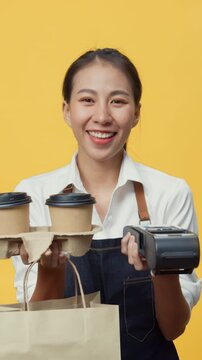 Vertical Screen : Happy smiling Asia waitress wear apron friendly looking at camera giving coffee or tea and credit card reader machine to customer  isolated on yellow background. Business takeaway.