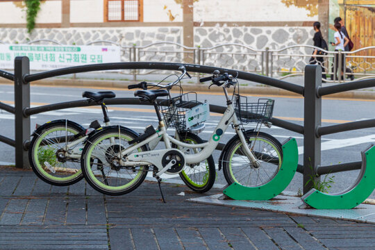 Green and white public rental bicycles are parked in a row at a sidewalk docking station with pedestrians in the background.