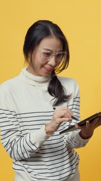 Vertical Screen : Happy Asia student lady holding tablet computer and pointing finger up at copy space enjoy looking book digital online shopping while standing isolated over yellow background.