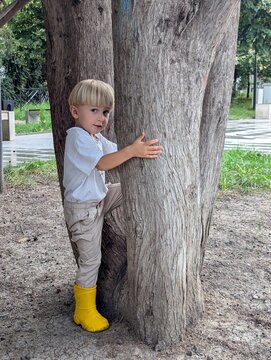 A young child in yellow rubber boots, crouched behind a large tree in the park. Quiet mischief and carefree summer moments.

