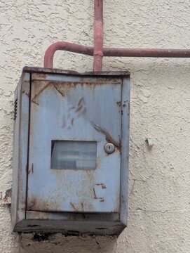 An old electricity meter hanging on a wall, supported by rusted pipes. Faded dial, peeling paint, and years of neglect.

