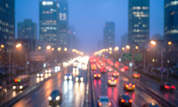 A blurred cityscape featuring busy traffic on a wet street during a rainy evening. Urban buildings and streetlights create a vibrant, atmospheric scene.