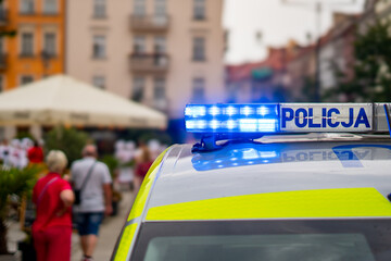 Police car roof close-up featuring an illuminated blue emergency light bar with POLICJA lettering, parked on a city square with blurred buildings, umbrellas, and pedestrians in daylight. © Adam