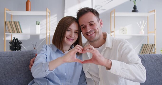 Couple form heart with hands at home. Smiling on couch in a bright livingroom, they express love and romance, creating a sweet gesture of affection indoors. Symbol of love and togetherness.