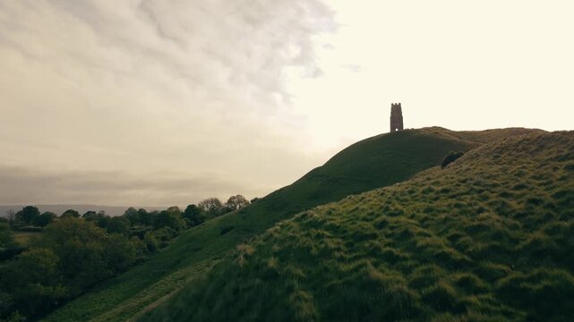 Aerial drone footage revealing a historic medieval tor with a tower, surrounded by lush green fields at sunrise.